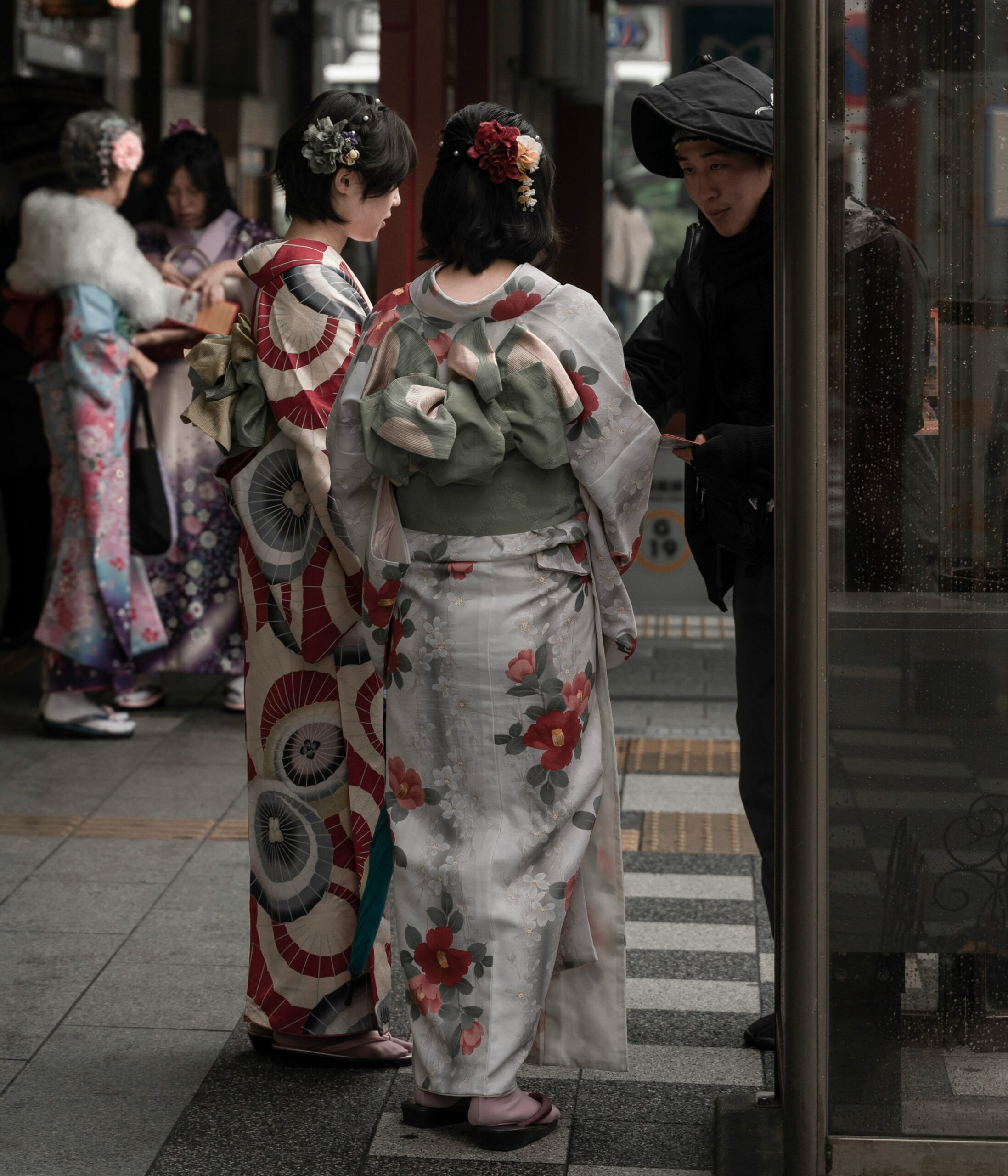 Two women in traditional kimonos with colorful patterns and floral hair ornaments seen from behind, engaged in conversation on a city street