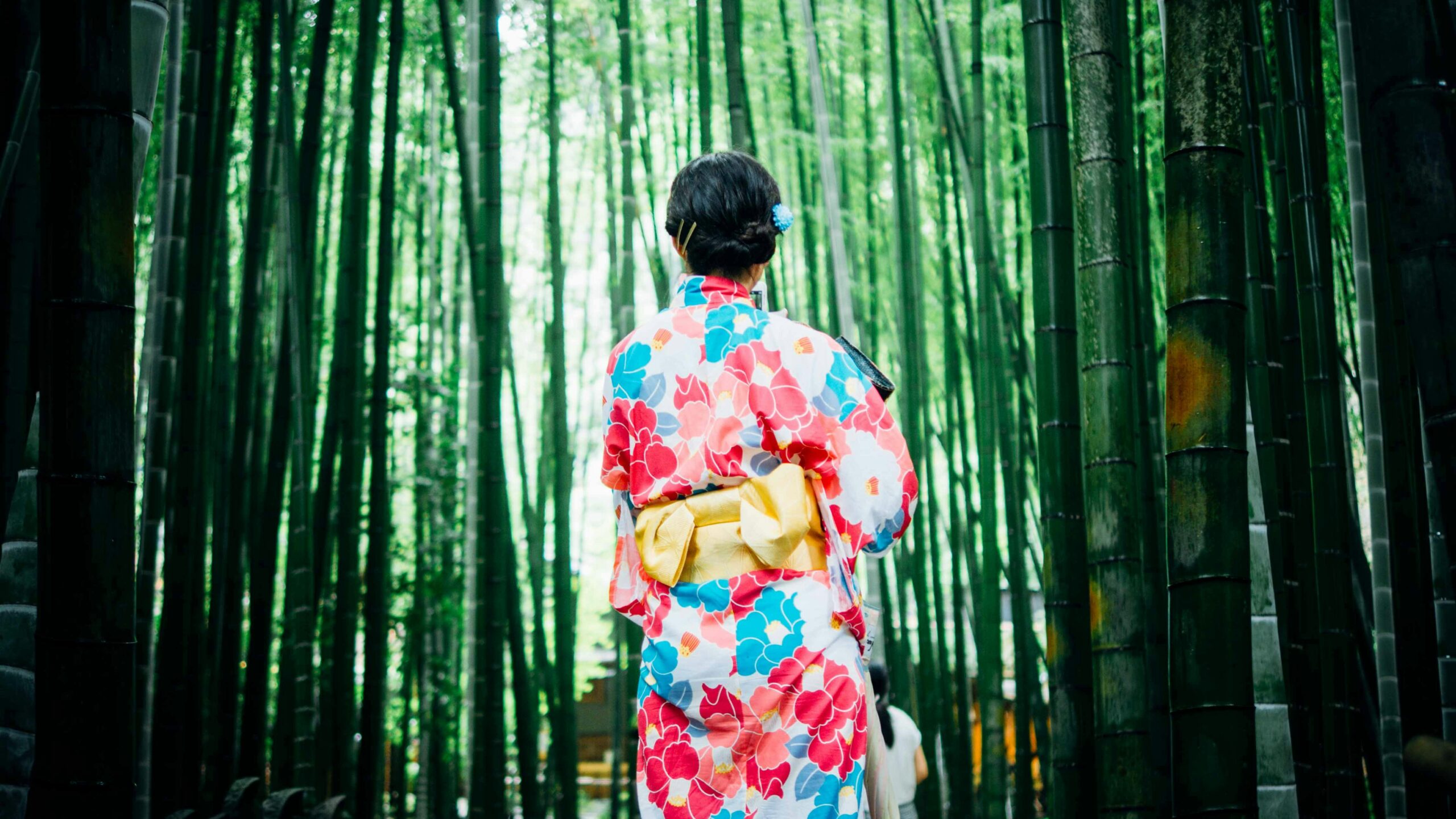 Person wearing a colorful floral kimono with yellow obi walking through a bamboo forest in Kyoto