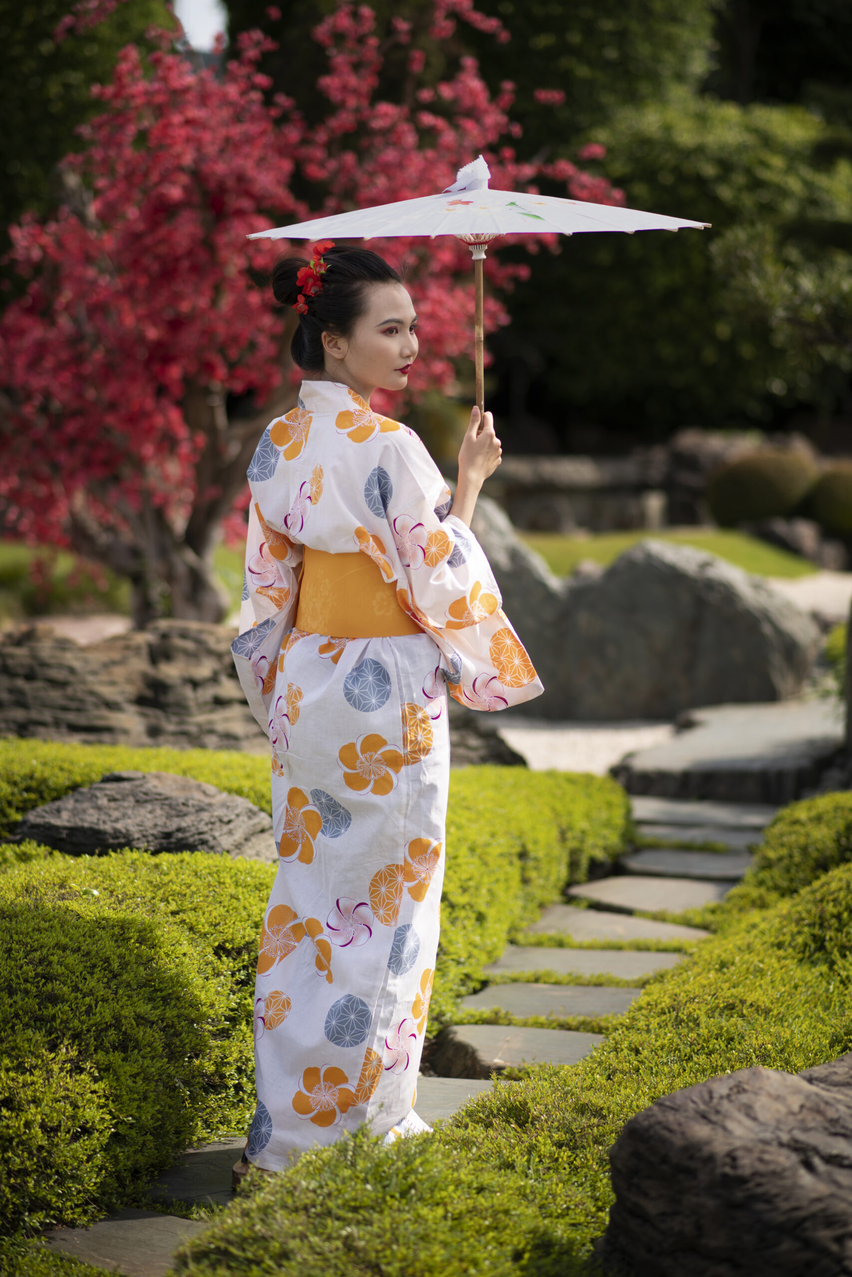 Person in a white kimono with orange floral patterns holding a parasol in a Japanese garden