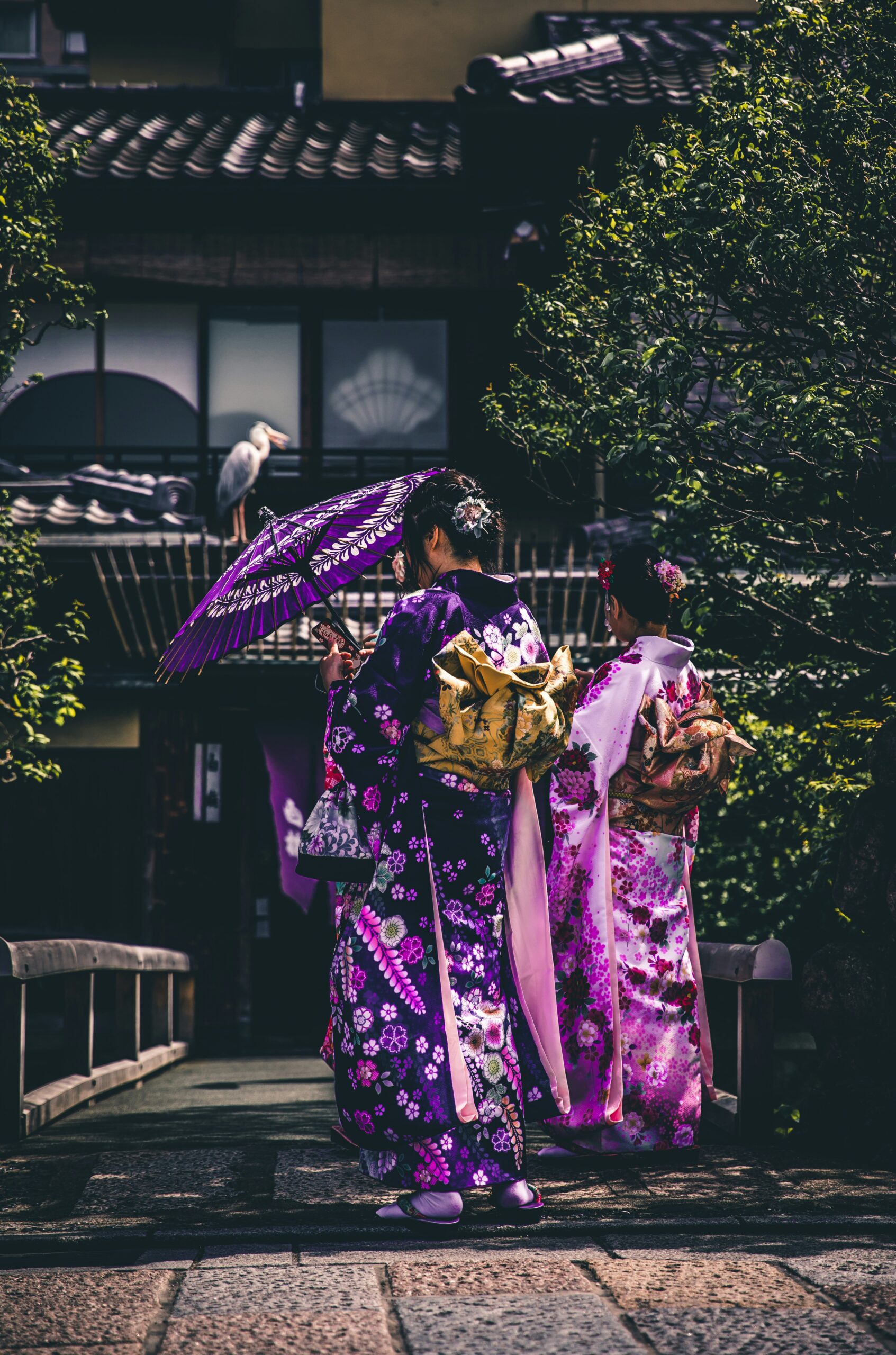 Two women in traditional Japanese kimonos with vibrant purple and pink floral patterns, one holding purple parasol, walking in scenic traditional street with wooden architecture and lush greenery