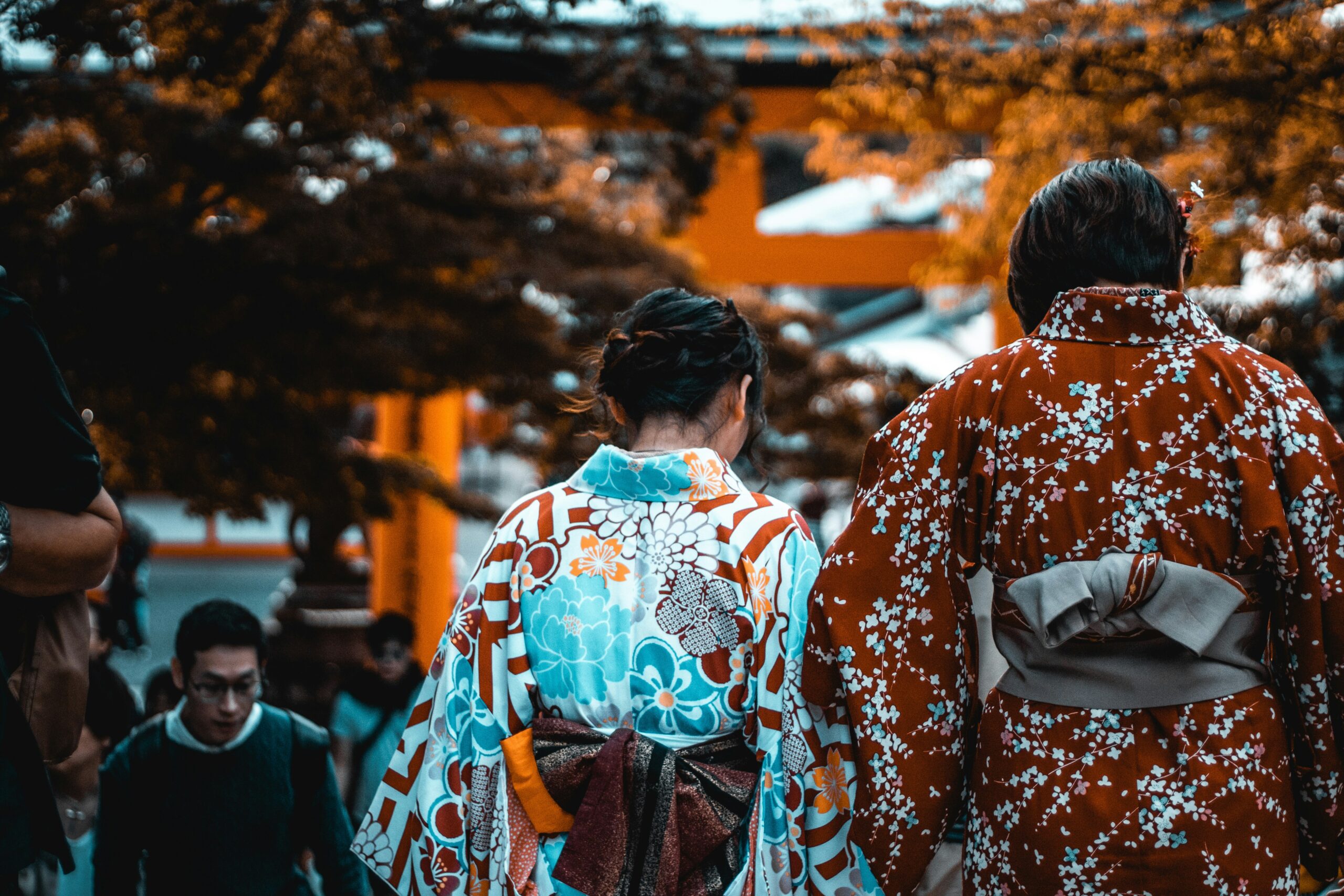 Modern Kyoto street scene in Gion district with people wearing beautiful kimonos walking past traditional wooden buildings, cherry blossoms, lanterns and historic architecture showing continuation of kimono culture in contemporary Kyoto