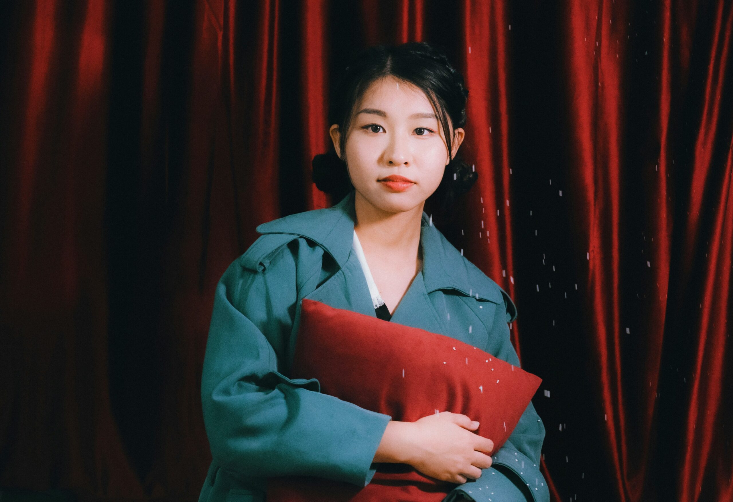 Young person in teal green coat holding red accessories against rich crimson curtain backdrop