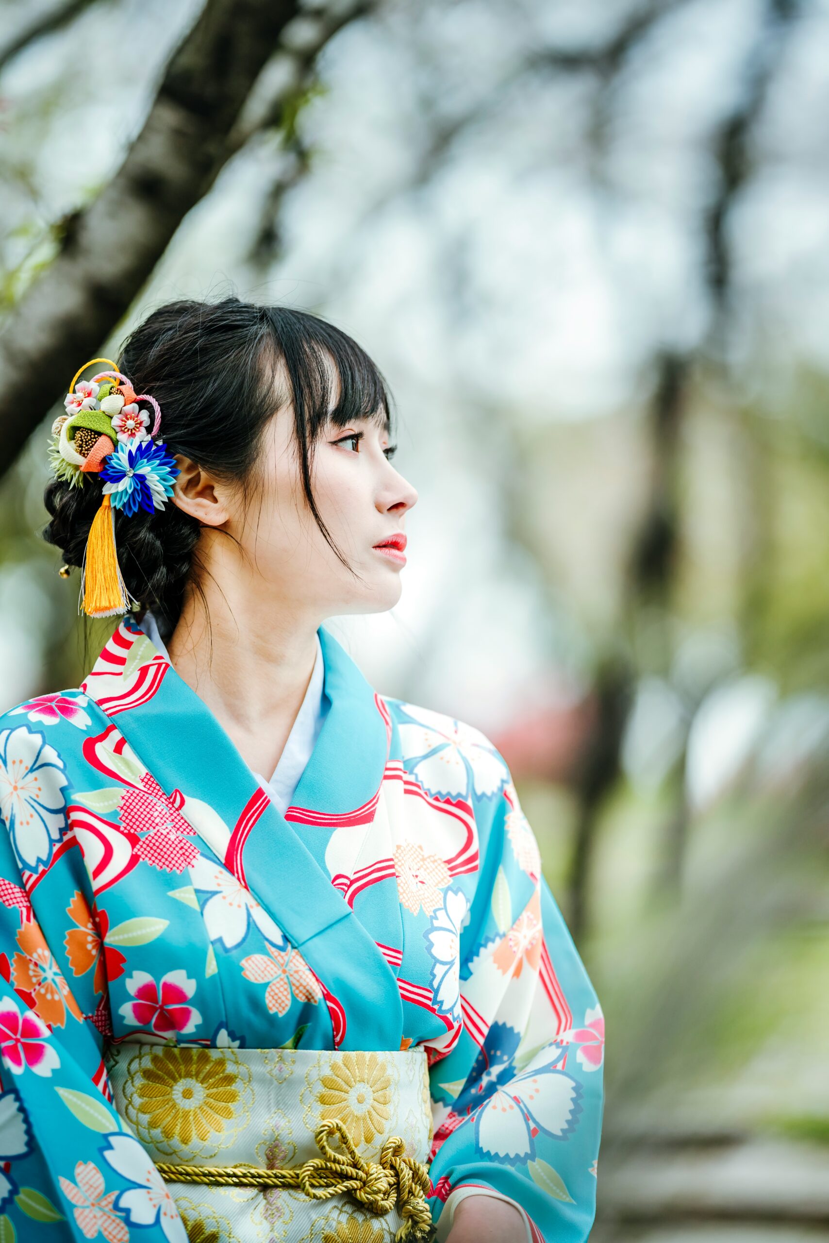 Mujer joven con un kimono turquesa brillante y adornos de pelo coloridos mirando hacia arriba en un entorno natural al aire libre, pose perfecta para un retrato de primavera.