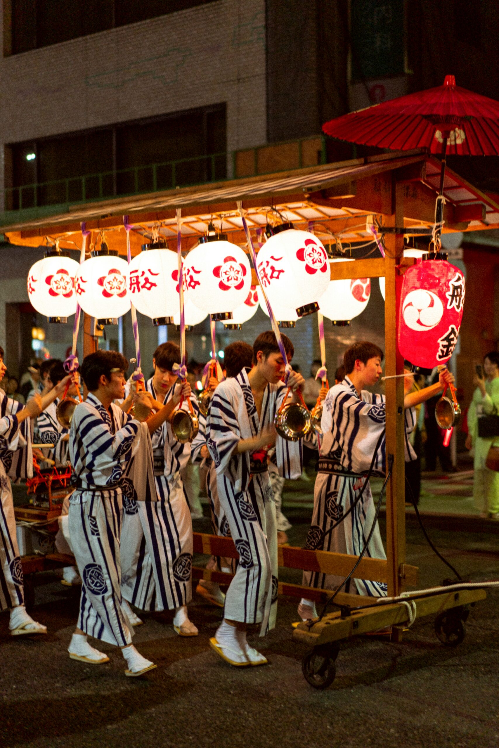 Japanese festival participants in traditional blue and white striped yukatas playing instruments while pulling festival float decorated with red and white paper lanterns with Japanese characters