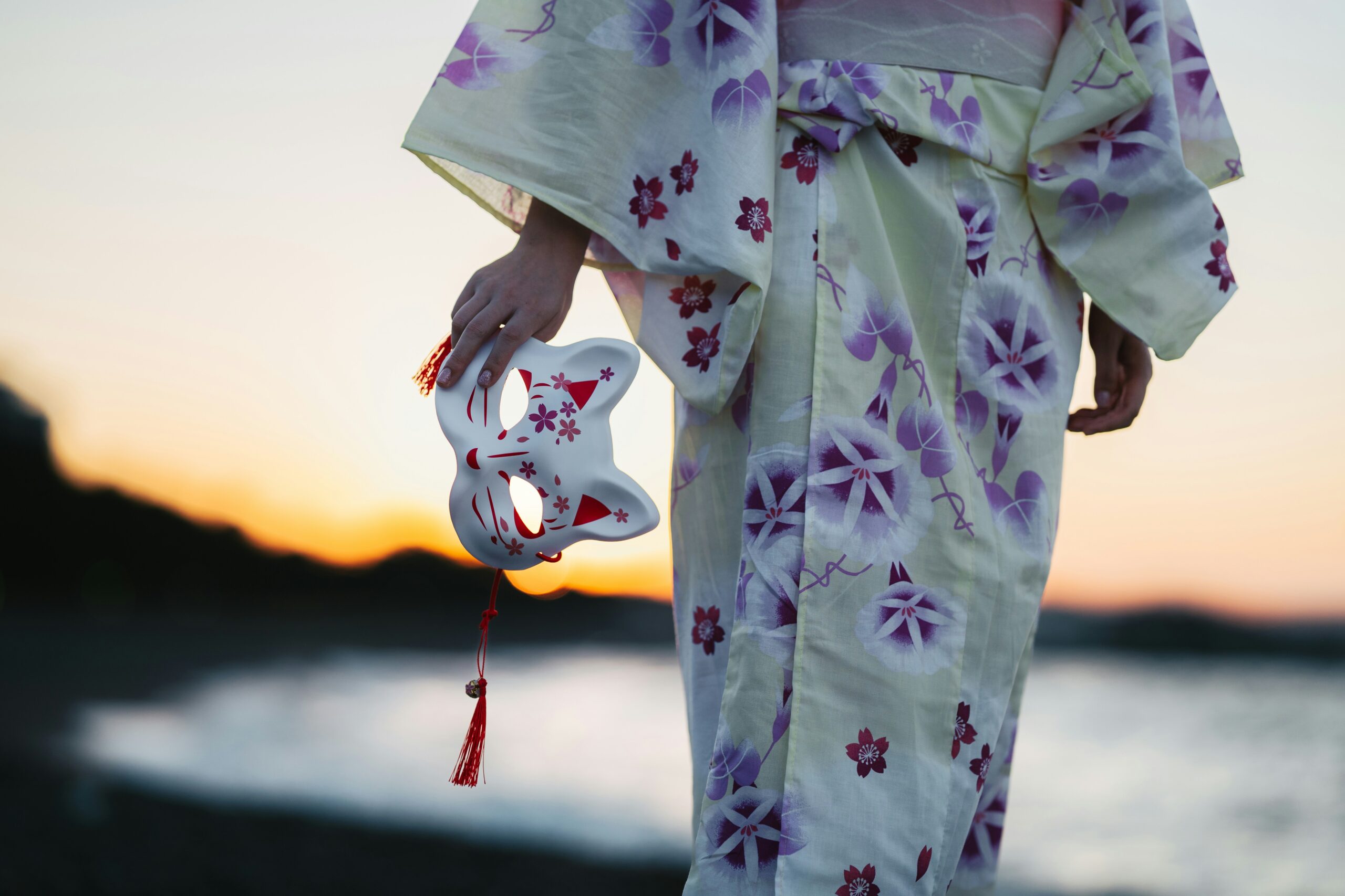 Woman in elegant floral kimono holding traditional Japanese mask at sunset over water, showcasing perfect kimono photography lighting and cultural authenticity in Kyoto