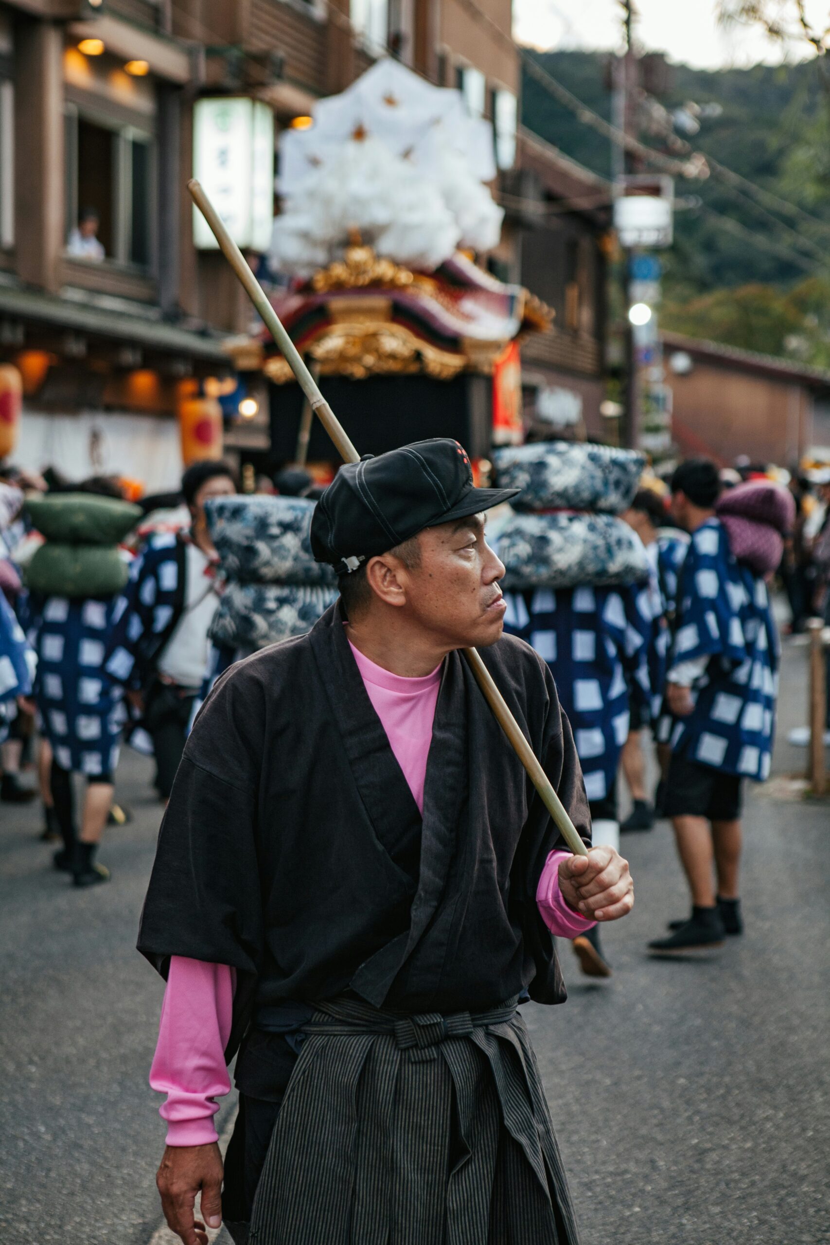 Traditional Japanese festival matsuri participant in dark robe and pink shirt holding bamboo stick during street celebration with portable shrine float in background